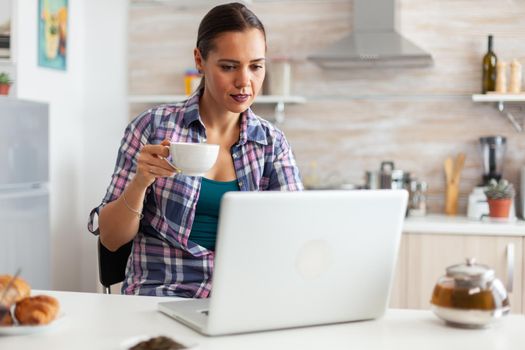 Woman holding cup of hot tea and using laptop in the morning. Working from home using device with internet technology, browsing, searching on gadget in the morning.