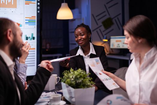 Workaholics focused multi-ethnic businesspeople overworking in company office meeting room brainstorming ideas late at night. Coworkers sitting at desk analyzing statistics paperwork