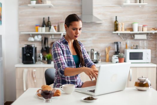 Lady pointing at laptop screen during morning in kitchen with breakfast next to her and cup of green tea. Working from home using device with internet technology, browsing, searching on gadget in the morning.