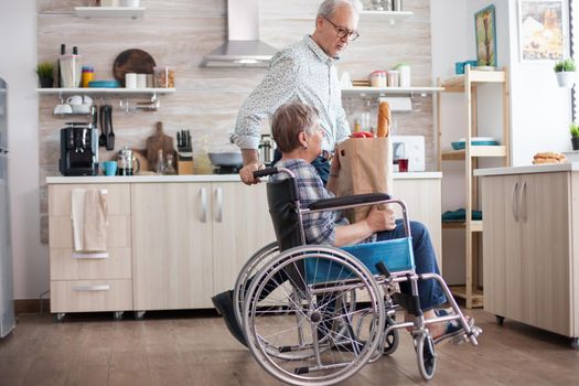 Senior man taking grocery paper bag from handicapped wife in wheelchair. Mature people with fresh vegetables from market. Living with disabled person with walking disabilities