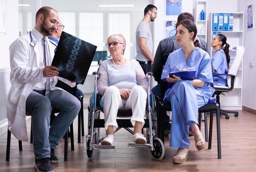 Doctor with stethoscope holding disabled senior woman radiography in wheelchair while talking with her in hospital waiting area. Patient asking about his appointment at clinic reception.