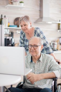 Disabled senior man in wheelchair and his wife having a video conference on laptop in kitchen. Paralyzed old man and his wife having a online conference.