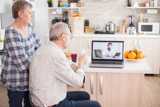 Senior man showing pills bottle to doctor while using laptop in kitchen for telemedicine. Online health consultation for elderly people drugs ilness advice on symptoms, physician telemedicine webcam. Medical care internet chat