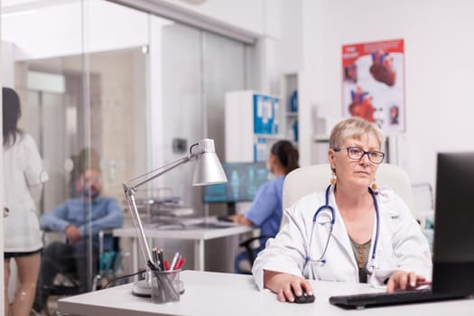 Senior woman doctor working on computer in hospital office while young medic is discussing with disabled patient in wheelchair on clinic corridor and nurse in blue uniform.