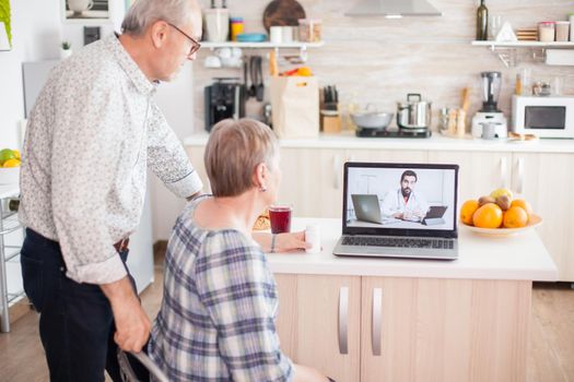 Doctor on telemedicine for senior couple. Video conference with doctor using laptop in kitchen. Online health consultation for elderly people drugs ilness advice on symptoms, physician telemedicine webcam. Medical care internet chat.