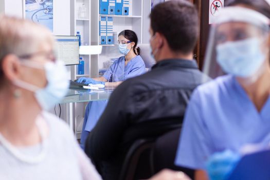 Medical staff working on computer in hospital hallway wearing face mask against coronavirus as safety precaution. Patient and medical staff in waiting area. Doctor in examination room.