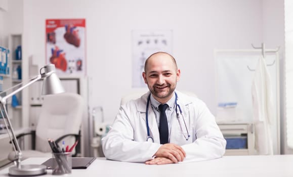 Portrait of successful young doctor in hospital office wearing white coat smiling at camera.