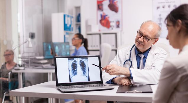 Senior doctor pointing at lungs x-ray on laptop for young patient in hospital office. Mature disabled woman in wheelchair discussing with medic about illness.