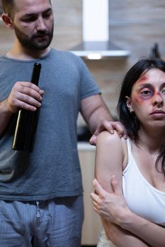 Alcoholic husband harassing abused wife with bruises. Violent man holding bottle of alcohol threatening terrified woman. Victim of domestic violence and physical trauma, marriage problem.