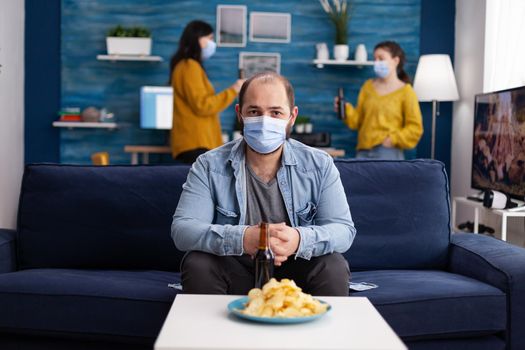 Young relaxed man wearing face mask looking at camera as safety prevention sitting on couch while speding time with friends during global pandemic with coronavirus. Group of young people having fun.