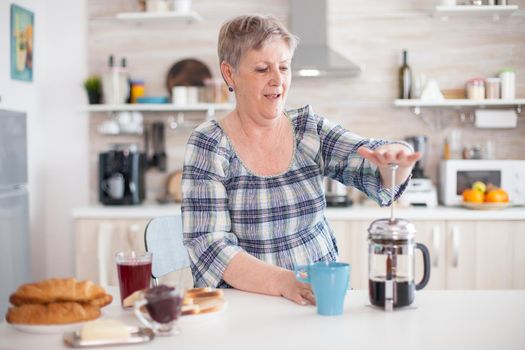 Mature woman using french press to make coffee in kitchen during breakfast. Elderly person in the morning enjoying fresh brown cafe espresso cup caffeine from vintage mug, filter relax refreshment