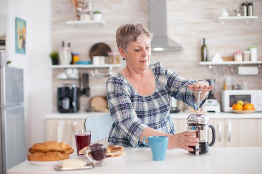 Senior woman making coffee with french press in kitchen during breakfast. Elderly person in the morning enjoying fresh brown cafe espresso cup caffeine from vintage mug, filter relax refreshment