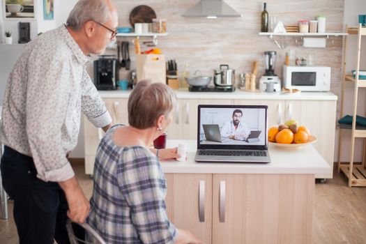 Senior couple during online medical check up from their home. Video conference with doctor using laptop in kitchen. Online health consultation for elderly people drugs ilness advice on symptoms, physician telemedicine webcam. Medical care internet chat