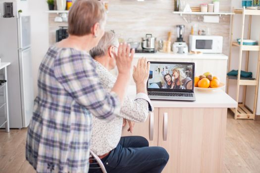 Happy senior woman waving during a video conference with family using laptop in kitchen. Online call with daughter and niece. Elderly person using modern communication online internet web techonolgy.