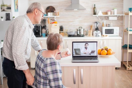 Mature woman talking with doctor via internet connection on her laptop. Video conference with doctor using laptop in kitchen. Online health consultation for elderly people drugs ilness advice on symptoms, physician telemedicine webcam. Medical care internet chat