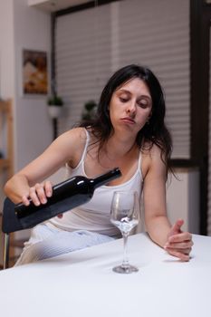 Young adult drinking wine alone in kitchen at home. Heartbroken woman pouring glass of alcohol from bottle having unhealthy addiction. Depressive person feeling miserable and intoxicated