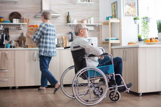 Invalid senior man smiling looking through the window in kitchen and wife is unpacking groceries. Invalid, pensioner, handicapped, paralysis.