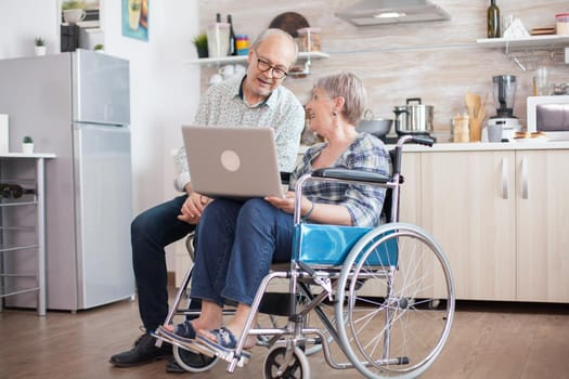 Disabled senior woman in wheelchair and her husband having a video conference on tablet pc in kitchen. Paralyzed old woman and her husband having a online conference.