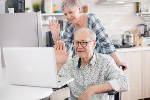 Invalid man and wife saying hello to their family. Disabled senior man in wheelchair and his wife having a video conference on laptop in kitchen. Paralyzed old man and his wife having a online conference. Invalid man and wife saying hello