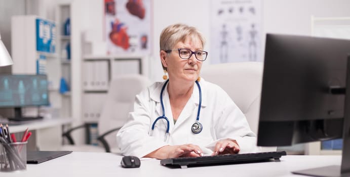 Mature female doctor using computer in hospital cabinet.