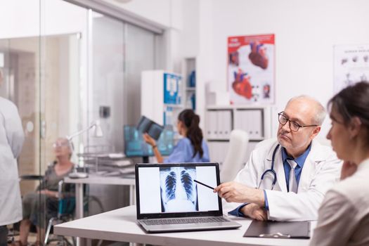 Senior doctor pointing at x-ray of healthy lungs on laptop in hospital office during patient medical visit. Young medic explaining diagnosis to disable mature woman in wheelchair.