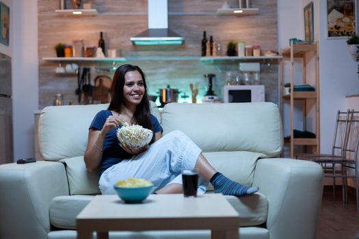 Woman laughing watching tv and eats snacks. Young happy, excited, amused, home alone lady enjoying the evening sitting on comfortable couch dressed in pajamas eating popcorn in front of television