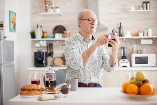 Pensioner paying online using credit card and application from smartphone during breakfast in kitchen. Retired elderly person using internet payment home bank buying with modern technology