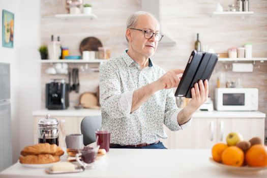 Happy elderly person with portable tablet PC in kitchen during breakfast. Senior person with tablet portable pad PC in retirement age using mobile apps, modern internet online information technology with touchscreen