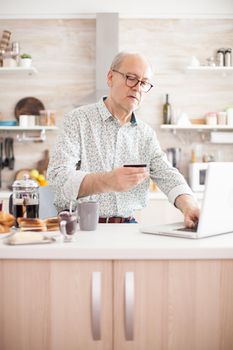Elderly man checking finances, holding a credit card in his hand. Pensioner paying online using credit card and application from laptop during breakfast in kitchen. Retired elderly person using internet payment home bank buying with modern technology