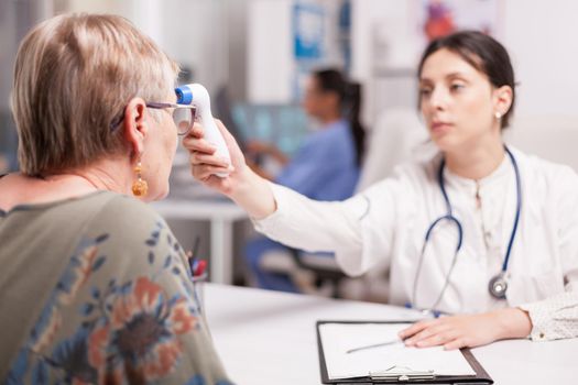 Medic using thermometer gun on senior patient during check up for infection with coronavirus. Doctor wearing white coat and stethoscope in hospital office.