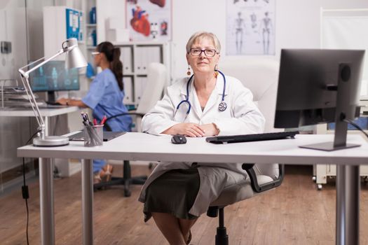 Senior medical therapist in hospital cabinet and nurse working on computer in the background.