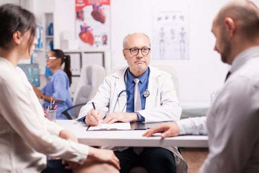 Senior doctor writing notes on clipboard in hospital cabinet during consultation of young couple. Medical practitioner explaining diagnosis. Nurse in blue uniform working on laptop.