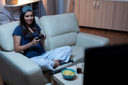 Woman sitting on sofa playing video game late at night wering eye mask on forehead. Excited determined gamer using controller joysticks keypad playstation gaming and having fun winning electronic game