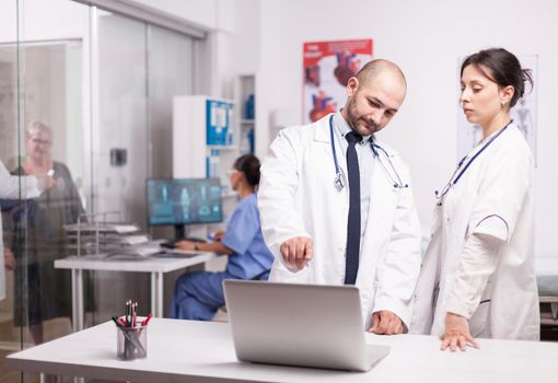 Young doctors pointing at laptop screen in hospital office dressed with white coat. Elderly aged woman and medic in clinic corridor talking about illness prescription. Nurse in blue uniform working on computer.