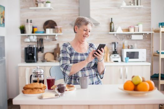 Old woman browsing on internet using smartphone in kitchen during breakfast. . Online communication connected to the world, senior leisure time with gadget at retirement age