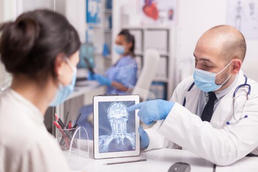 Doctor with face mask against coronavirus showing patient brain trauma on tablet computer. Medic wearing white coat and stethoscope.