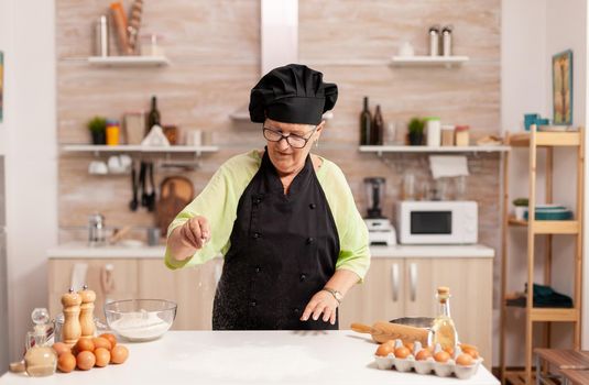 Woman preparing homemade cookies spreading flour table in home kitchen. Happy elderly chef with uniform sprinkling, sieving sifting raw ingredients by hand baking homemade pizza