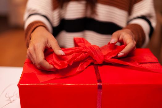 Close up of present box wrapping red ribbon for bow in kitchen with decorations, ornaments, lights and tree. Young caucasian woman decorating gift for christmas eve night celebration