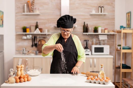 Senior lady chef spreading flour with hand for food preparation in home kitchen wearing apron. Happy elderly chef with uniform sprinkling, sieving sifting raw ingredients by hand baking homemade pizza.