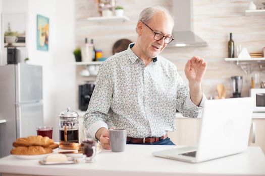 Cheerful senior man on online video call from kitchen while enjoying breakfast and a cup of coffee. Elderly person using internet online chat technology video webcam making a video call connection camera.