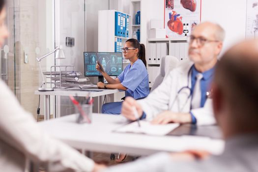 Nurse in blue uniform holding x-ray image in hospital cabinet. Older doctor wearing white coat during consultation of disabled man in wheelchair and his wife.