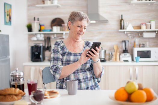 Senior woman using mobile gadget in the kitchen. Authentic elderly person using modern smartphone internet technology. Online communication connected to the world, senior leisure time with gadget