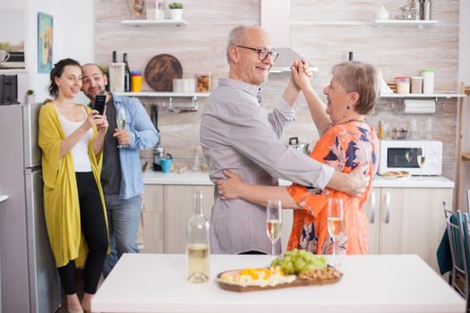 Happy senior couple dancing in kitchen during family celebration. Delicious assorted cheese on wooden board. Woman taking photos.