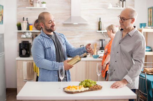 Son holding wine bottle in kitchen while having a conversation with his senior old. Wfe and mother preparing delicous lunch for family meeting.