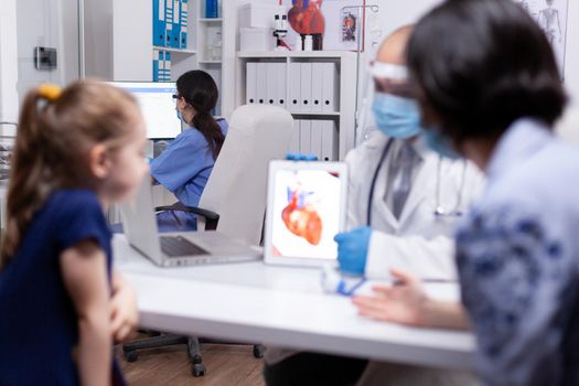 Physician talking about heart functions with patients during coronavirus using tablet. Pediatrician doctor in protection gloves and mask providing clinic medicinal health care services.