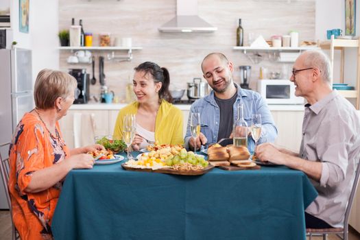 Mother and daughter smiling while having delicious lunch in kitchen with family. Senior man holding wine glass. Tasty roasted potatoes.