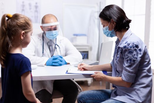 Mother writing child treatment on clipboard sitting in medical office. Health pediatrician specialist providing health care services consultations treatment in protective equipment for consultation