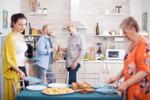 Father and son toasting with wine and smiling at each other. Mother and daughter setting plates on table for lunch.