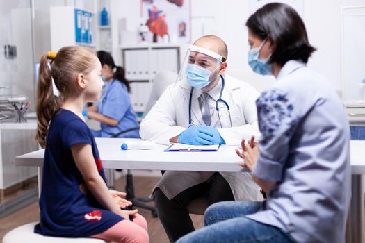 Physician having conversation with child during consultation and coronavirus outbreak in hospital office wearing face mask. Health pediatrician specialist providing health care services consultations treatment.