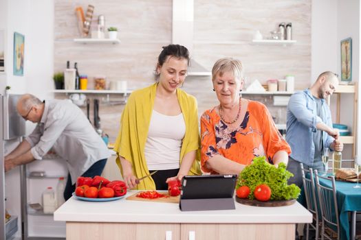 Senior woman watching salad recipe on table while cooking together with her daughter in kitchen. Mother daughter preparing lunch.